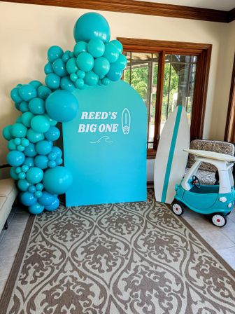 Turquoise balloon arch and matching backdrop with surfboard prop and toy ride-on car in a sunroom — seaside-themed first birthday photo display