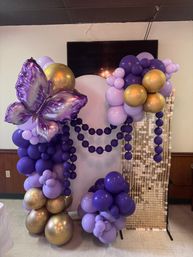 Purple and gold balloon arch with large iridescent purple butterfly foil balloon, lavender and deep purple clusters, and sequin photo backdrop for indoor party event
