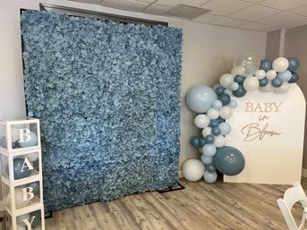 Indoor baby shower backdrop featuring a textured blue flower wall, stacked clear blocks spelling BABY with balloons, and a blue-and-white balloon garland framing a "Baby in Bloom" sign on a wood-look floor