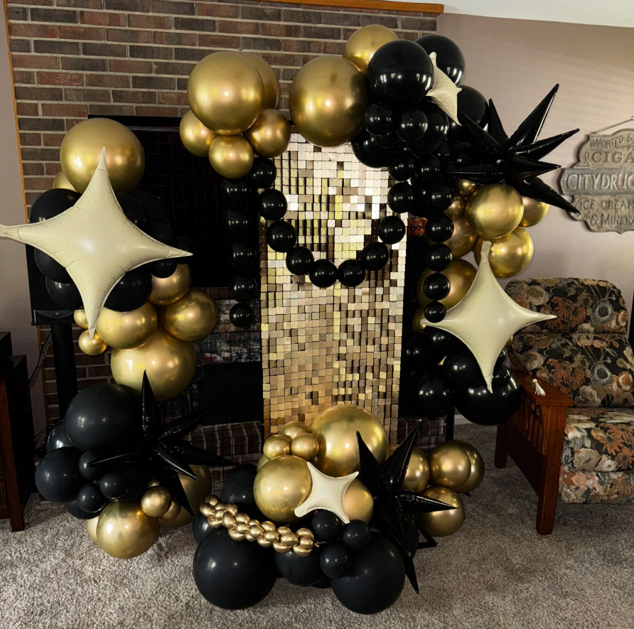 Glam gold-and-black balloon arch with star-shaped and metallic balloons framing a gold sequin photo backdrop in a cozy living-room party setup.