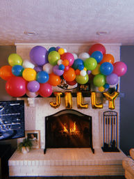 Vibrant multicolored balloon garland draped over a white brick fireplace mantel in a living room, warm fire glowing behind a black screen and shiny gold foil letter balloons below — cozy indoor party decor.