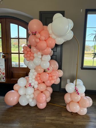 Peach and white balloon garland with small pink floral accents on a gold hoop stand framing a wooden front door and window — indoor party or event decor at a home's entryway