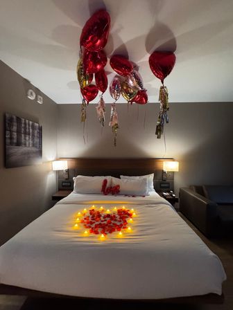 Romantic hotel room interior with a king bed decorated with a heart-shaped ring of glowing tealight candles and red rose petals, red foil heart balloons floating at the ceiling and a 'love' pillow on the headboard.