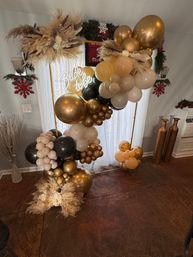 Glam indoor bridal shower backdrop: gold, black and cream balloon arch with pampas grass and a glowing "Bride to be" sign in a cozy living room setting