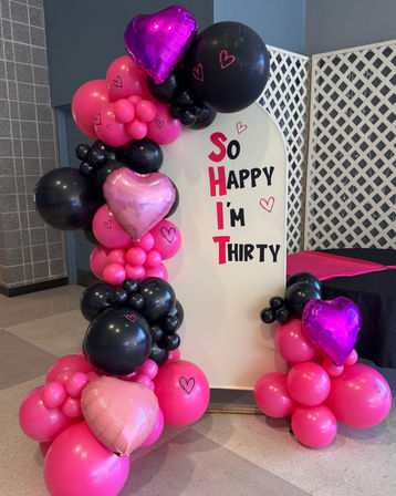 Playful pink and black balloon arch with purple and pink heart foil balloons framing a white sign that reads 'So SHIT I'm Thirty' — 30th birthday party decor in an indoor event space.