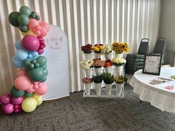 Cheery indoor event setup with a colorful balloon arch framing a white backdrop that reads "She's off the Market", a rack of white buckets filled with sunflowers and mixed blooms, and a round table with a 'Flower Bar' sign.