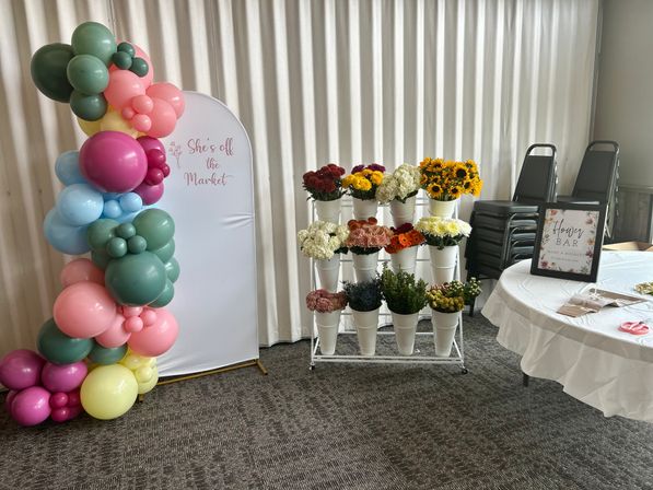Cheery indoor event setup with a colorful balloon arch framing a white backdrop that reads "She's off the Market", a rack of white buckets filled with sunflowers and mixed blooms, and a round table with a 'Flower Bar' sign.