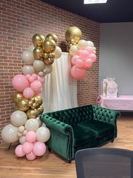 Emerald green velvet tufted sofa against an exposed brick wall and cream curtain, styled with a pink, gold and ivory balloon arch over a wooden floor and nearby gift table for a celebration backdrop.