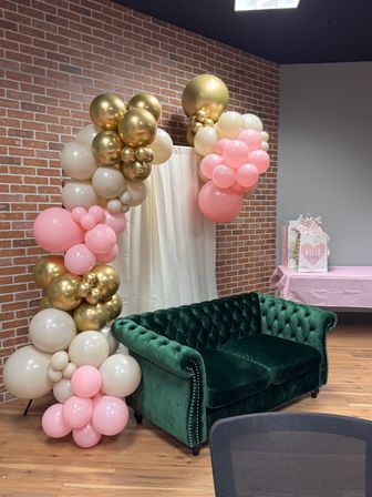 Emerald green velvet tufted sofa against an exposed brick wall and cream curtain, styled with a pink, gold and ivory balloon arch over a wooden floor and nearby gift table for a celebration backdrop.