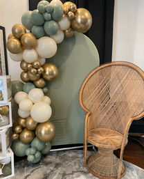 Boho event photo backdrop: sage-green, cream, and metallic gold balloon garland arching around a round sage panel beside a woven rattan peacock chair on a neutral rug