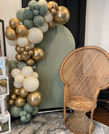 Boho event photo backdrop: sage-green, cream, and metallic gold balloon garland arching around a round sage panel beside a woven rattan peacock chair on a neutral rug