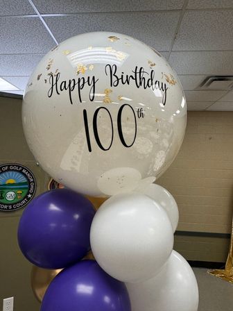 Clear oversized balloon with gold confetti reading Happy Birthday 100th, paired with purple, white and gold balloons in an indoor event space