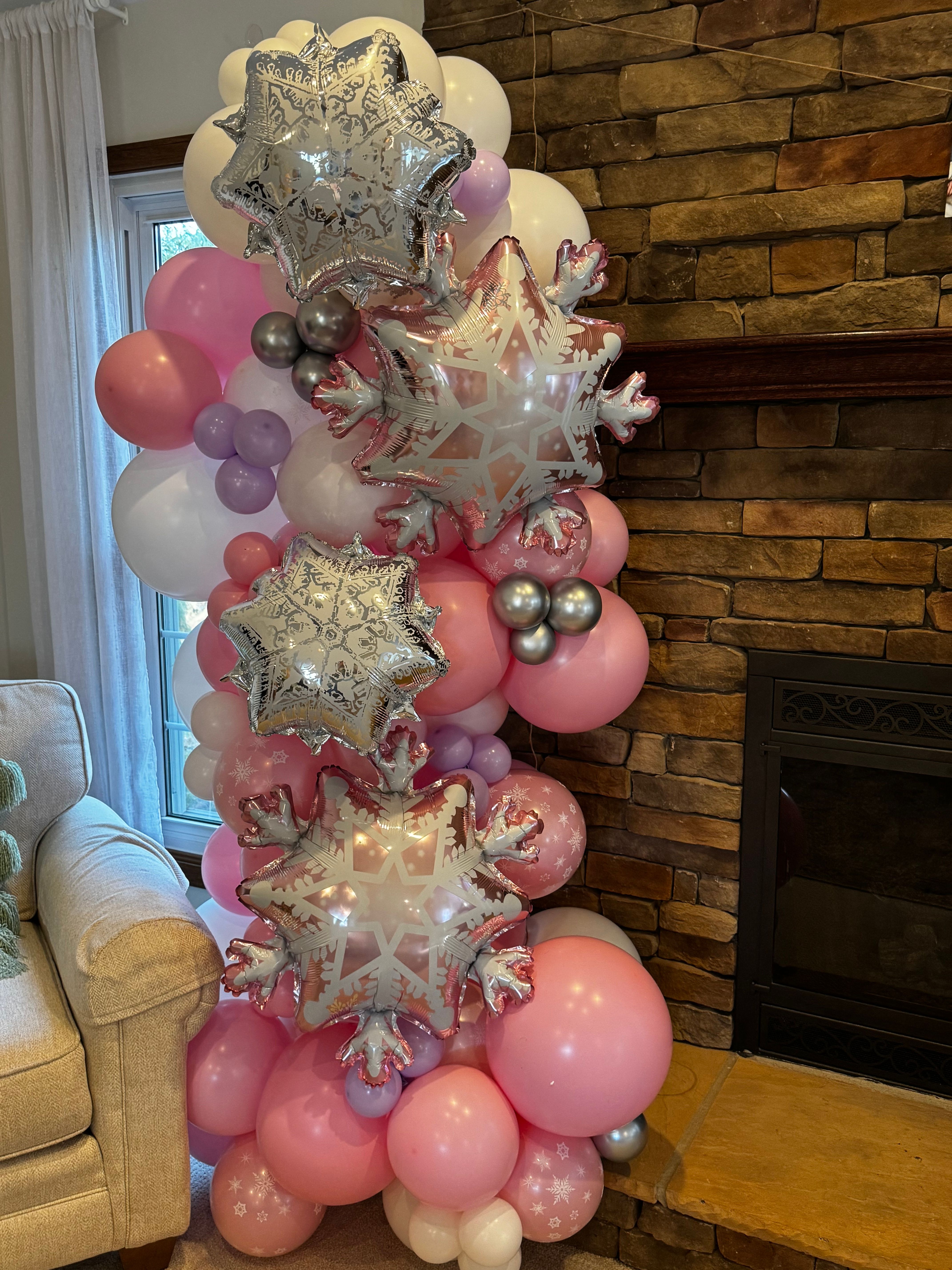 Indoor living room balloon column of pink, lavender and white latex balloons accented with silver snowflake star foil balloons staged beside a stone fireplace and window