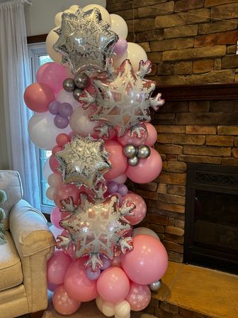 Indoor living room balloon column of pink, lavender and white latex balloons accented with silver snowflake star foil balloons staged beside a stone fireplace and window