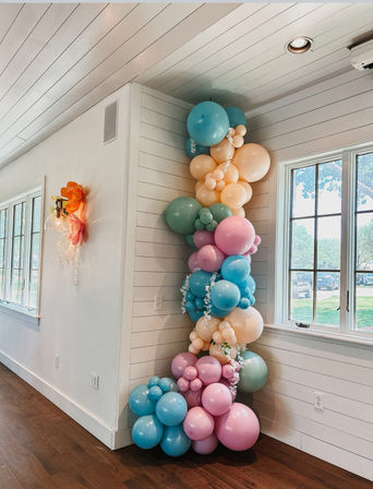 Playful pastel balloon garland column in a bright farmhouse-style room — clustered pink, blue, peach and mint balloons along a white shiplap corner next to a window over hardwood floors, perfect for indoor party decor.