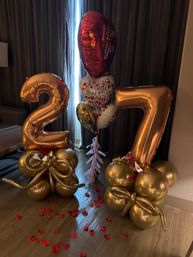 Romantic indoor balloon arrangement featuring rose-gold foil numbers '27', heart-shaped "I Love You" balloons, gold balloon clusters and scattered red rose petals on a wooden floor.