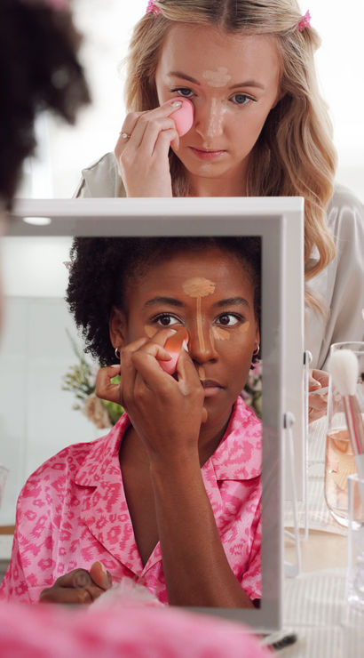 Two women applying concealer with pink beauty blenders at a vanity mirror during a playful morning makeup routine
