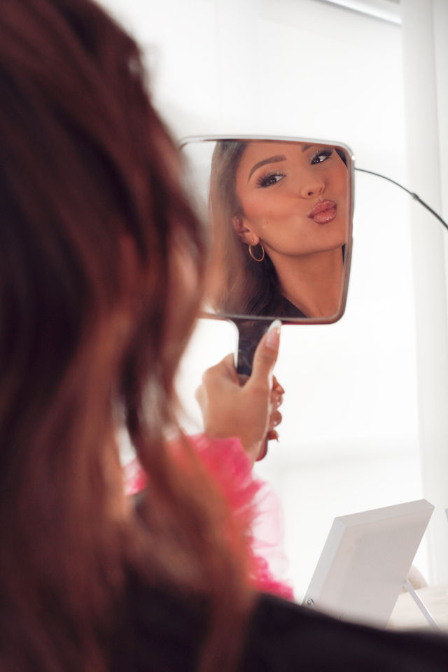 Woman checking glam makeup in a handheld mirror, puckering glossy lips and admiring long lashes at a bright home vanity