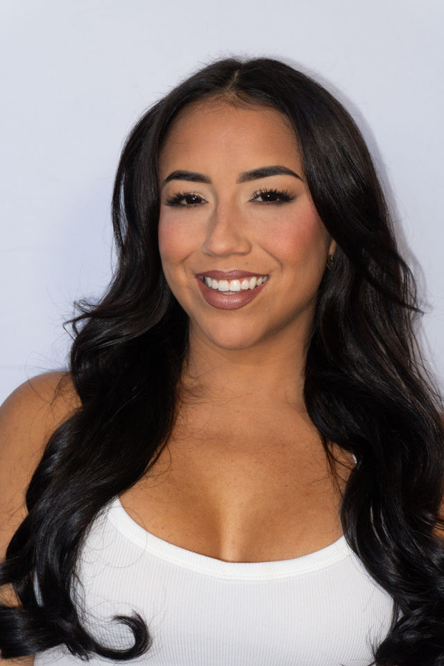 Close-up studio portrait of a smiling woman with long dark wavy hair, full lashes and warm makeup, wearing a white tank top against a light background.