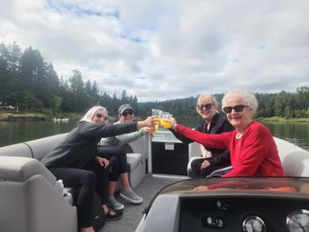 Four friends wearing sunglasses toasting with orange drinks on a pontoon boat on a calm lake, with a forested shoreline and cloudy sky in the background.