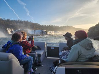 Four bundled passengers and a dog on a pontoon boat viewing a wide cascading waterfall and mist over a forested river at sunset