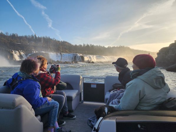 Four bundled passengers and a dog on a pontoon boat viewing a wide cascading waterfall and mist over a forested river at sunset