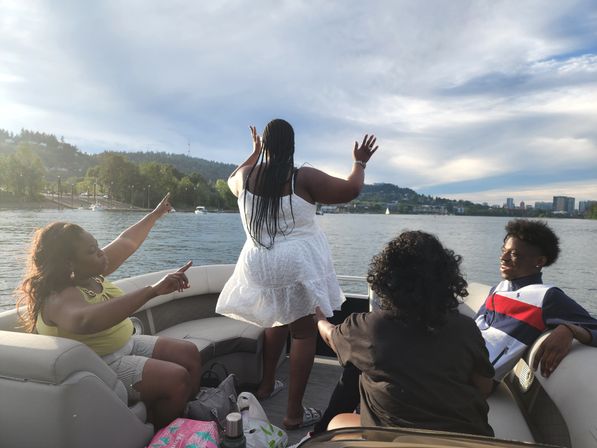 Four people enjoying a pontoon boat ride on a calm river near an urban riverfront; one person in a white sundress stands at the bow with arms raised while others sit, point and laugh under a soft late-afternoon sky.