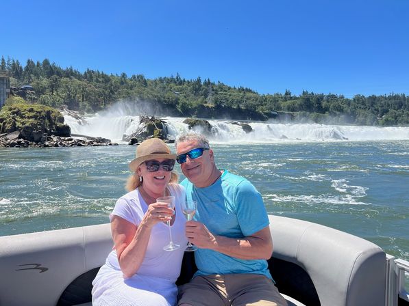 Couple toasting with wine on a boat in front of a wide cascading river waterfall and forested shoreline on a sunny summer day