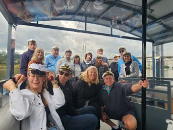 Smiling adults wearing captain hats on a covered riverboat cruise, posing under a cloudy sky with a cable-stayed bridge and urban riverfront in the background.