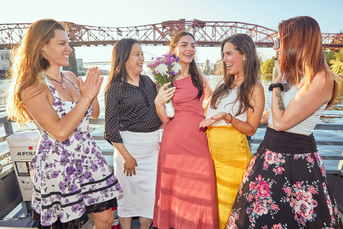 Group of five women celebrating on a riverboat, one holding a bouquet, with a red steel bridge and urban waterfront in the background.