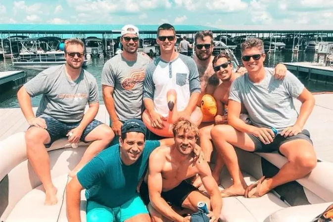 Eight friends posing on a pontoon boat at a marina on a sunny summer day, some in swim trunks and sunglasses, one holding a pink inflatable flamingo and others holding drinks with covered boat slips and calm water in the background.