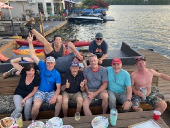 Nine friends laughing and posing on a lakeside dock during a playful summer gathering, with kayaks, boats and picnic plates and drinks visible on the waterfront.