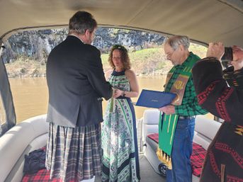 Couple exchanges rings during intimate pontoon boat wedding on a river; groom in a kilt, bride in a green patterned dress, officiant reads from a book while a guest photographs.