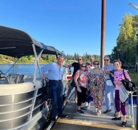 Group of adults smiling on a sunny lakeside dock next to a pontoon boat, wearing colorful summer clothes and sunglasses with a forested shoreline and clear blue sky