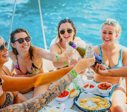 Friends in swimsuits on a boat over bright blue water, clinking champagne flutes and toasting above a spread of chips, salsa and snacks on a sunny boat party.