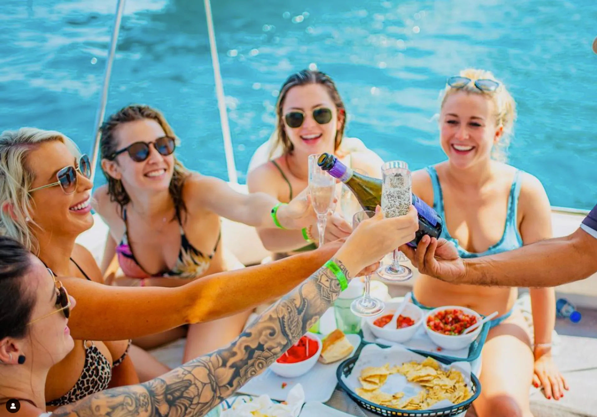 Friends in swimsuits on a boat over bright blue water, clinking champagne flutes and toasting above a spread of chips, salsa and snacks on a sunny boat party.