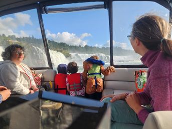 Family on a sightseeing boat tour watching a large cascading waterfall through the windows, kids in life jackets leaning on the railing with mist and blue sky.