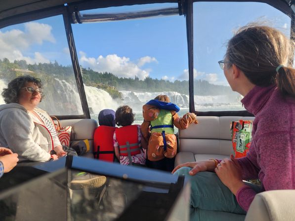 Family on a sightseeing boat tour watching a large cascading waterfall through the windows, kids in life jackets leaning on the railing with mist and blue sky.