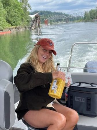 Smiling person in a red cap on a pontoon boat pouring a drink from a yellow bottle, cruising a tree-lined river with a steel bridge in the distance.