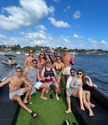 Group of friends on a sunny boat party at a palm-lined waterfront inlet, posing on a yacht deck with artificial turf in swimwear and colorful drinks