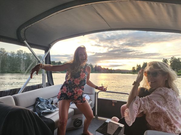 Two friends laughing on a pontoon boat at sunset on a tree-lined lake, one dancing in a floral romper holding a drink and the other seated with sunglasses in a relaxed summer boating scene