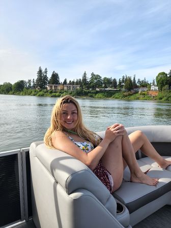 Smiling woman lounging on a pontoon boat seat on a sunny summer day, with a calm river and tree‑lined waterfront homes along the shore in the background.