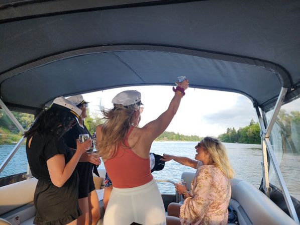 Group of women in white captain hats raising drinks aboard a pontoon boat cruising a tree-lined lake, summer celebration vibe.