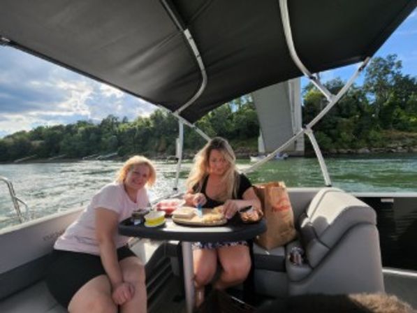 Two people on a pontoon boat under a black canopy preparing sandwiches at a small table, cruising a tree-lined river beneath a concrete bridge on a sunny afternoon.