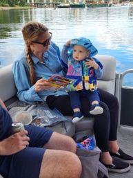 Woman in sunglasses reads a colorful picture book to a baby in a blue sun hat and life jacket while they sit on a pontoon boat on a calm lake.