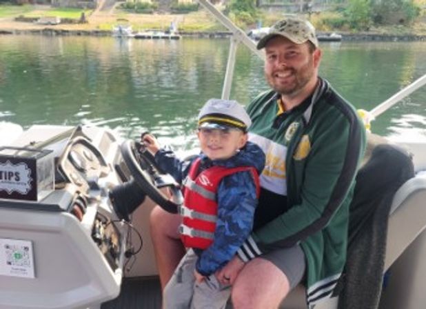 Man and young child smiling on a lake boat; child wearing a captain’s hat and red life jacket at the steering wheel, calm water and tree-lined shore in the background.