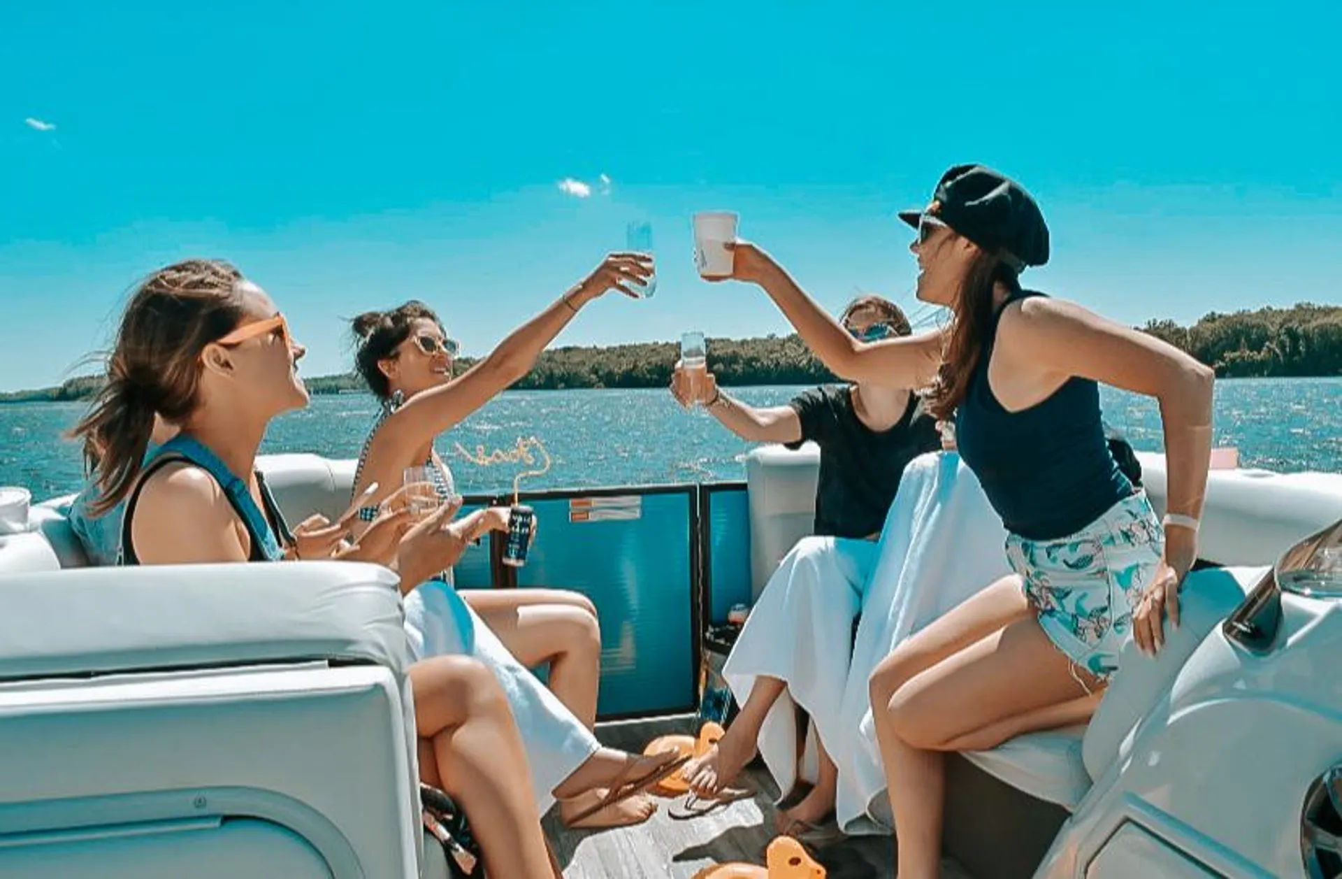 Four friends clinking drinks on a sunny pontoon boat cruising a blue lake with a tree-lined shore — fun summer outing with sunglasses and swimsuits.
