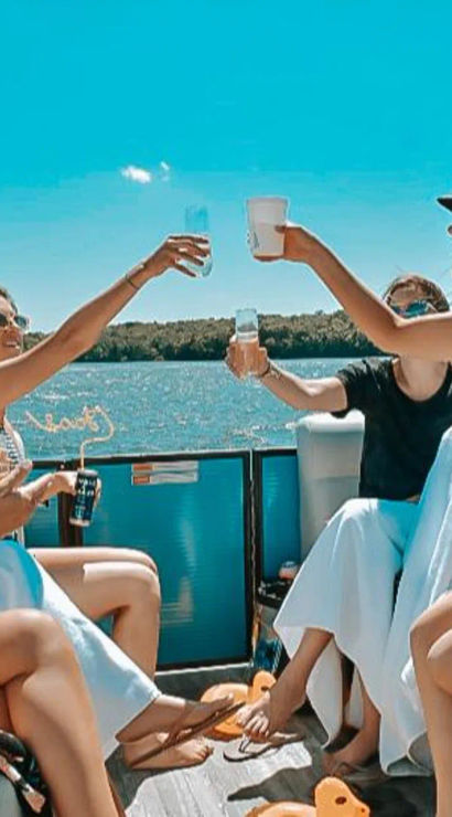 Four friends clinking drinks on a sunny pontoon boat cruising a blue lake with a tree-lined shore — fun summer outing with sunglasses and swimsuits.