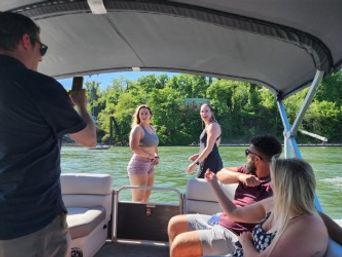Friends on a covered pontoon boat enjoying a sunny summer lake outing, two people on the swim platform and three seated with tree-lined shoreline in the background.