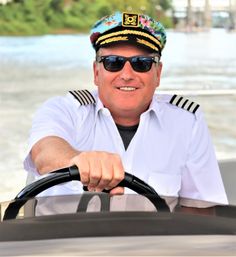 Smiling boat captain in a floral captain’s hat and sunglasses steering a motorboat on a sunny river waterway